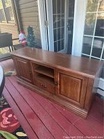 Wooden TV stand placed outdoors on a deck, showing full front view with two cabinets, central open shelves, and a drawer.