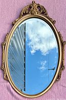 Front view of oval antique mirror with gilded ornate wood frame, top decorative crest visible.