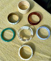 Eight vintage costume jewelry bangles displayed on a light fabric surface under natural light, showing different colors, materials, and styles.