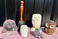 Front view of six owl figurines showing variety in color, material, and style including black glass, resin, wood, and clay-like pieces.