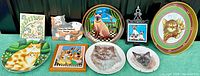 Full view of assortment of nine cat-themed plates and plaques arranged on green background showing various realistic and stylized cat depictions in different shapes and frames