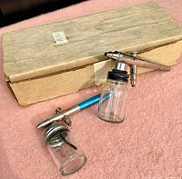 Two vintage airbrushes with attached glass paint jars placed on a fabric surface along with original cardboard box