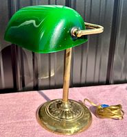Vintage green bankers desk lamp shown with green glass shade and brass base on a pink surface with a corrugated black background.