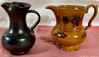 Two vintage English ceramic pitchers on pink cloth background, a dark brown Prinknash pottery pitcher and a tan Elijah Cotton Lord Nelson Ware pitcher with floral designs