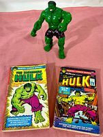 Photo of the Hulk action figure standing behind two vintage Incredible Hulk comic paperbacks on a pink surface, showing the cover art and figure details.