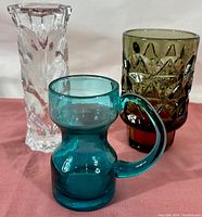Lot includes three vintage Mid-Century Modern glass items against a neutral background: a blue glass pitcher with handle, a heavy clear pressed glass candlestick and a smoked geometric patterned glass vase.