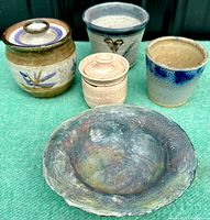Six vintage handcrafted pottery pieces displayed on a turquoise surface showing various styles, glazes, and lidded and open vessels.