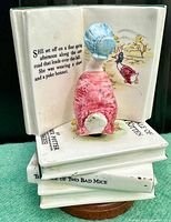 Front view of the ceramic music box showing the seated girl figure in a red dress and blue poke bonnet facing an open book with text and illustration, atop three stacked ceramic books.