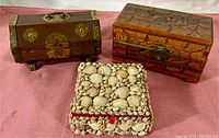 Three jewelry boxes on a pink fabric, a curved brass accented wood box, a rectangular carved wood box, and a square shell-covered box