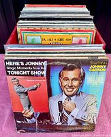 Photo of vintage vinyl records in a red crate, focusing on the front album with Johnny Carson on the cover from Here's Johnny: Magic Moments from the Tonight Show.
