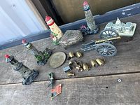 Photo showing metal miniature lighthouses, small cannon, lead soldier figurine, brass vessels, a metal decorative box, and a miniature American flag all displayed on a wooden surface.