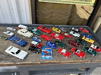 Wide view of approximately 20 diecast cars including vintage, convertible, and sports models on a wooden surface outdoors.