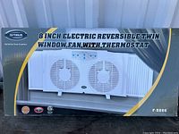 Front view of the boxed Optimus 8 inch electric reversible twin window fan with thermostat, showing the product image and branding.