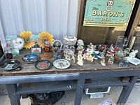 Wide view of the lot arranged on a wooden table outside showing various figurines, plates, sunflower wall art, and glass items.