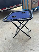 Side angle view of folding camping table showing blue fabric top with cup holders and black metal frame legs