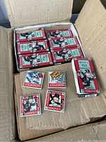 Open cardboard box showing multiple boxed sets of Monopoly porcelain tile magnets. Each box labeled 'Set of 4 Tile Magnets' with Mr. Monopoly image. Several individual magnets displayed outside boxes showing designs including Mr. Monopoly face and game tokens such as '500' money and dice.