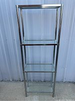 Front view of the glass shelf with metal frame showing three clear glass shelves, standing against an outdoor wall.