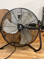 Front view of HomeRitz floor fan showing circular grille, three metal blades, black center hub with logo, and supporting black frame on wooden surface.