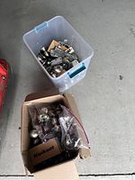 Top view of plastic container with mixed door knobs, hinges, and hardware pieces inside next to a box with additional parts and plastic bags.