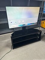 Samsung 55 inch smart LED TV turned on showing TV source menu mounted on a black TV stand with three shelves. Power cord visible on floor.