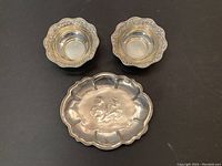 Three Indian silver bowls arranged on a dark surface showing two smaller bowls with decorated rims and one larger bowl with central floral motif.