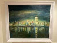 Overall view of the largest cityscape painting in white frame showing dark moody background with building reflections in water below.