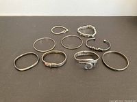 Nine sterling silver bracelets in a variety of designs including bangles, cuffs, chain link, and buckle style set on a plain surface.