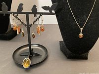 Full view of 3 pairs of amber earrings, sterling silver ring with amber, and sterling silver chain with amber pendant on black display stands.