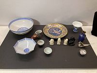 Wide shot showing all Asian collectible items including bowls, small figurines, decorative plate, vase, and small cups arranged on black surface.