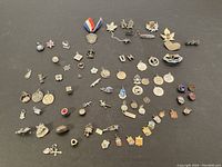 Wide shot showing an assortment of silver charms and pins arranged on a dark surface, displaying various shapes including hearts, crosses, medals, and decorative pins with small colored stones and enamel detail.