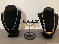 Front view of malachite necklace on left bust, multi-stone necklace on right bust, orange bead earrings on bird stand center.