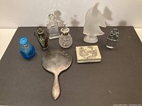 Front view of assorted perfume bottles, a frosted glass fish-shaped bottle, a decorative hinged box, and a sterling handheld mirror laid out on a black surface.