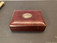 Closed rectangular jewelry box made of burgundy Italian leather with visible light wear and scuffs on the surface. Top center has an oval-shaped sterling silver badge with hallmarks and slight tarnishing.