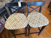 Two black wooden bar stools with beige and light brown patterned cushioned seats, X-back design visible, on hardwood floor.