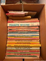 Top-down view of a box filled with about 26 vintage Peanuts and Charlie Brown books by Charles M. Schulz, showing various titles stacked horizontally.