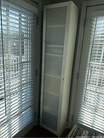 Tall white IKEA Billy bookcase with frosted glass door closed, positioned in a bright room corner near windows with blinds.