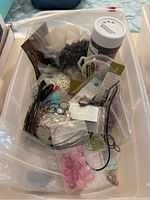 Top-down photo of a white plastic bin with assorted jewelry making supplies including beads, hooks, stones, cords, and small parts in bags and loose.
