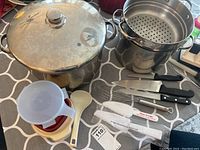 Photo showing two large stainless steel pots: one with fitted lid and one with perforated strainer insert. Three kitchen knives with black handles and a metal knife sharpener labeled 'Piece of Cake'. Several plastic containers of Tupperware and a white plastic serving spoon are also visible.