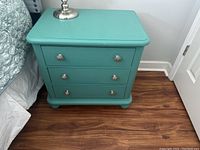 Photo of one aqua-colored nightstand next to bed, showing three drawers with round metal knobs on wood floor.