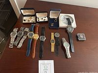 Wide view of nine men's wristwatches, three sets of cuff links in their boxes, and a small boxed collectible on a wooden surface.
