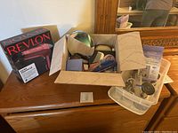 Photo of a dresser with a Revlon hair dryer box, open cardboard box with various bathroom and travel items, and transparent plastic container with toiletries.