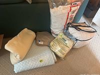 Photo showing folded off white blanket, tan sheets, white bag containing pillow, and packaged fillings.