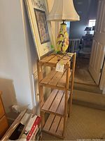 Photo showing vintage ceramic lamp with white fabric shade on top of wooden storage shelf next to framed art print leaning against wall.