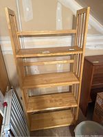 Light-colored wooden folding bookshelf with four shelves, slatted side panels, positioned against an unfinished wall.
