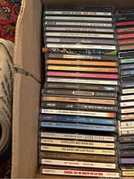 Photo showing a diverse collection of CD cases stacked in a box, featuring various artists and music genres.