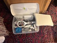 Open plastic container filled with white Nintendo Wii system components and accessories including three Wii Remotes, two Nunchuks, Balance Board, and Wii Sports game disc.