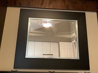 Photo showing large square mirror with thick black frame hanging on wall, reflecting part of a room with white wainscoting and door.