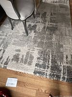 Photo showing a grey and off white area rug carpet placed on a wooden floor, with a chair partially visible on the rug. The rug has an abstract block pattern.