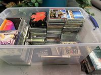 Large plastic tub filled with approximately 80 CDs of various artists, some still sealed in original plastic wrap, showing assorted album and compilation covers, providing a diverse music collection.