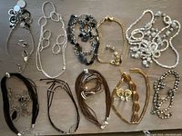 Top-down view of assorted necklaces, bracelets, and clip-on earrings laid flat on a wooden surface showing color and material variations including gold-tone metal, black and brown leather strands, and pearl-like beads.
